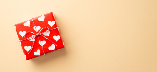 Valentine's Day concept. Top view photo of giftbox in wrapping paper with heart pattern and twine bow on isolated pastel beige background with copyspace