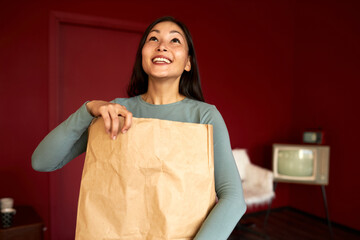 Fototapeta na wymiar Delivery service concept. Happy excited asian female holding paper bag with food or goods in hands, looking up with anticipation to open package, standing in stylish living-room with red walls