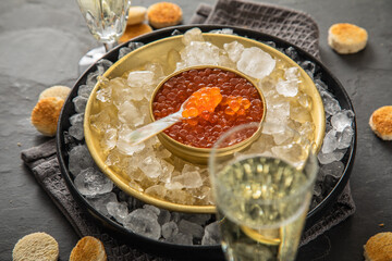 Orange Keta Lachs Kaviar in Dose auf Eis in Glas Schale mit Perlmutt Löffel, Sekt Champagner und Toast Brot auf dunkel Serviette und schwarz Schiefer