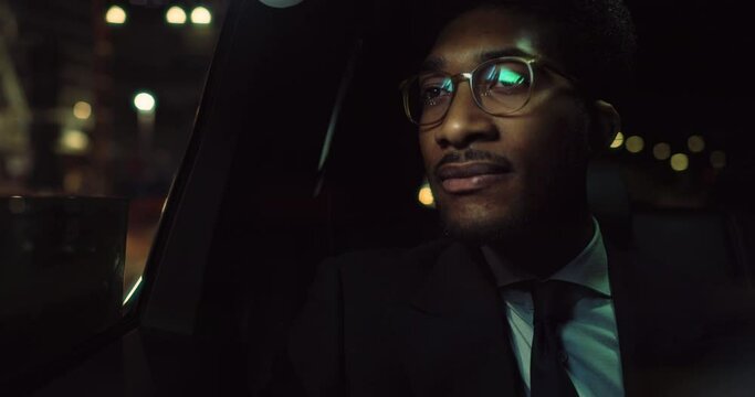 Portrait of Stylish Black Man in Glasses is Commuting Home in a Backseat of a Taxi at Night. Business CEO Looking Out of Window and Thinking while in a Car in Urban City Street with Warm Lights 