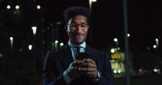 Closeup of a Handsome Stylish Black Male with Glasses Standing in a Street at Night, Using a Smartphone and Smiling. Portrait of an African American Man Doing Online Shopping, Chatting, Browzing