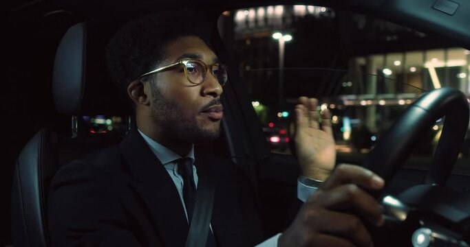 A Handsome Black Man in a Suit Driving a Fancy Car and Talking and Laughing on the Phone Remotely. Successful African American Businessman Having a Business Meeting with Investors While Driving
