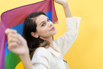 Happy cheerful caucasian white woman smiling and holding a rainbow flag which is the symbolic of LGBTQ people on yellow background. 