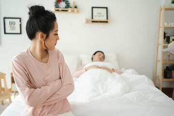 Obraz na płótnie Canvas Unhappy - stressful Asian woman sitting and crying on the bed while her husband sleeping, a family and post marriage problem. Arguing Asian young couple with bad - negative relationship.