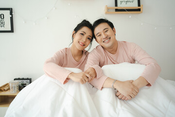 Fototapeta premium Happy and romantic Asian couple's portrait in bedroom with natural light from window, concept of relationship between husband and wife and being a family.