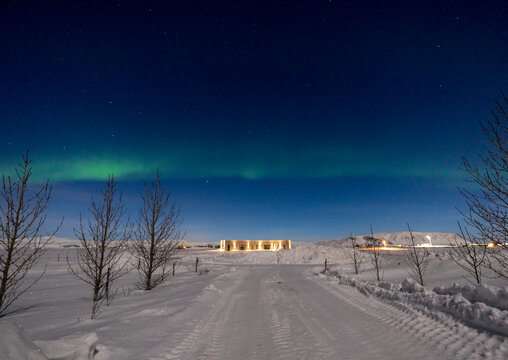 Northern lights on the horizon line and stars with a state of the art illuminated house at the end of a totally snowy road illuminated by moonlight in Iceland with tree silhouettes.