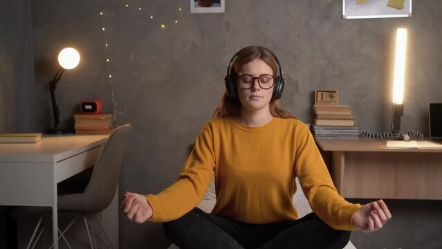A student girl sits in a college dormitory on a bed in a lotus position in an orange sweater with glasses, listens to music on headphones and meditates.