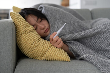 Sick Asian girl lying on sofa, wrapped in blanket and holds thermometer to measure body temperature. Unhealthy Korean woman infected with new strain of flu needs hospitalization. New covid-19 outbreak - obrazy, fototapety, plakaty