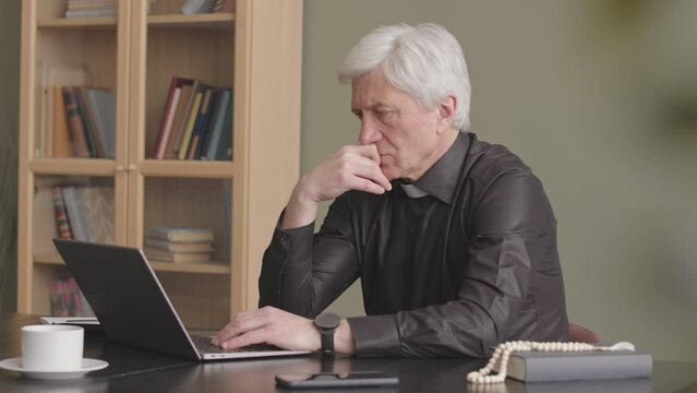 Pensive senior grey haired priest in black shirt and clerical collar working on laptop from home