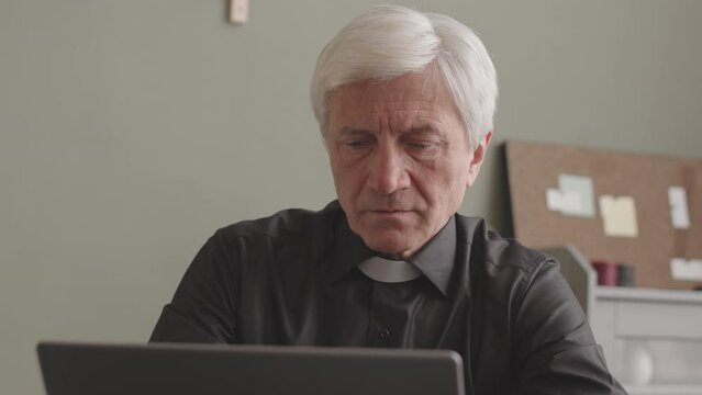 Thoughtful senior Caucasian priest in black shirt and clerical collar touching his chin while working on laptop from home office