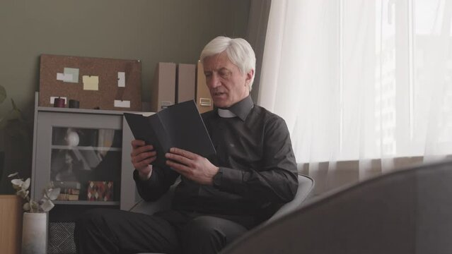 Senior priest in black shirt and white clerical collar reading Bible aloud at home