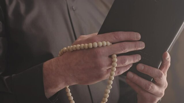 Slowmo of mature grey haired priest in black shirt and white clerical collar reading prayer indoors holding Bible and rosary beads in hands