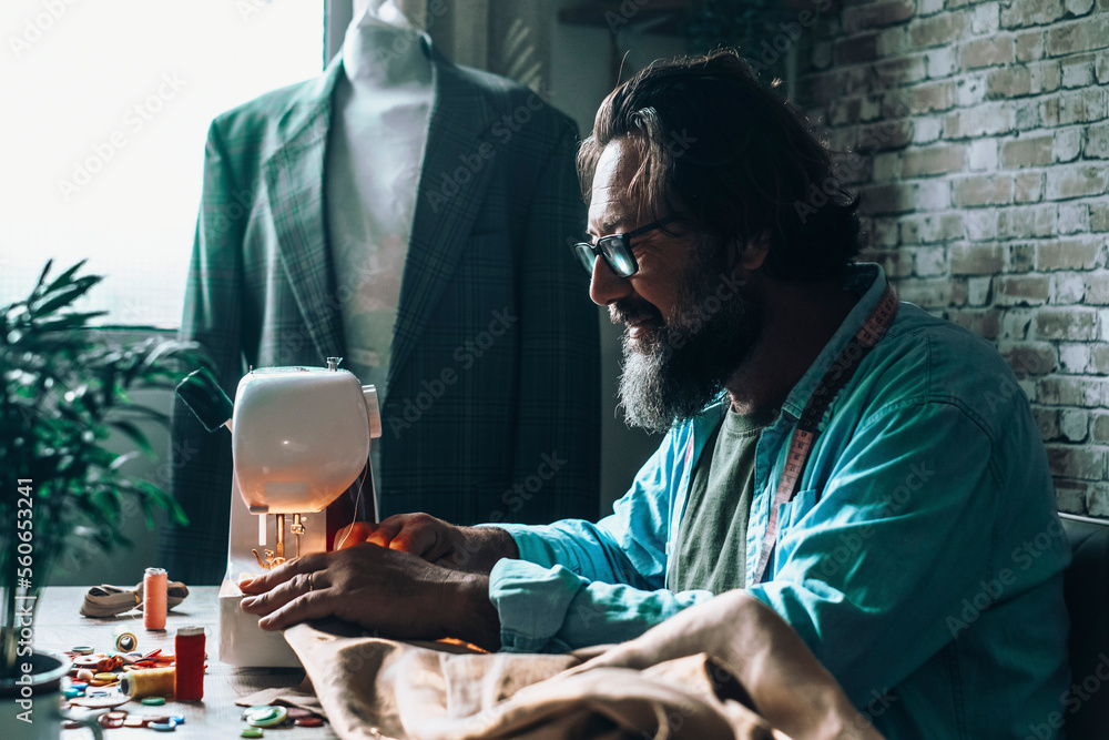 Wall mural Tailor man working on sewing machine in workshop with dummy wearing suit in background. Mature happy man work indoor and smile. Concept of alternative job activity foe male people with beard