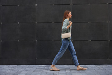 Young woman in casual clothes walking near grey wall outdoors, space for text