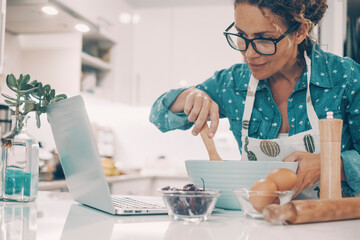 Housewife at work in the kitchen preparing bakery recipe listening and watching content on line in cooking channel. Modern woman cooking with online help. Laptop and people at home in domestic life.
