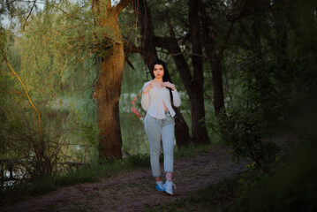 Fototapeta na wymiar Sexy sensual brunette walks in a summer forest at sunset in a white blouse and jeans