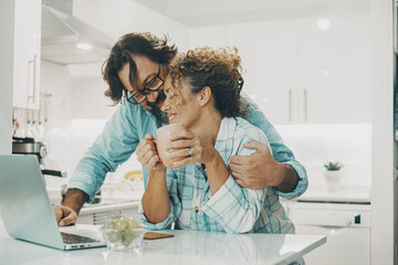 Fototapeta premium Man hugging woman at home in the kitchen with love. Indoor leisure activity and relationship lifestyle. Married people enjoying time. Young couple using computer and have romantic moment in real life