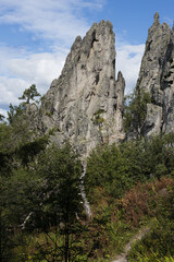 Majestic sharp grey rock as jagged pillar with cracks surrounded green trees, bushes in bright sunny day with white clouds in blue sky and sunlights, closeup, vertical. Amazing trekking background.