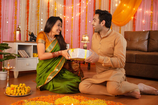 Young Indian couple celebrating Diwali festival by exchanging gifts - surprised wife. Husband and wife wearing traditional clothes sitting together and exchanging gifts - celebrating Diwali at home...