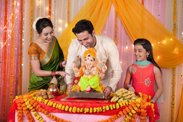 Cheerful Indian family welcoming Lord Ganesha idol on Ganesh festival / Ganesh Chaturthi. Smiling and happy family welcoming Lord Ganesh idol for Ganesh Chaturthi - Table decorated with marigold fl...