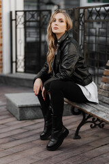 Fashionable  blonde woman model with  black leather jacket and style sunglasses sitting on a bench at the city 
