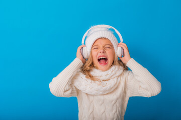 portrait of a happy little blonde girl in a white hat and sweater listening to music with headphones on a blue background. Winter, space for text