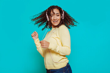 Young arabic smiling woman girl moves her curly hair in the air, wears yellow sweater, listens mp3 music podcast with pink headphones at studio isolated over blue background.