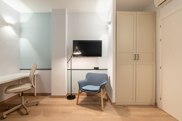 Interior of room with white walls, wardrobe, an entrance door and an air conditioner above it. Blue lounge chair bright lights and TV on wall, table and leather office chair for online remote work.