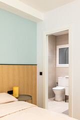 Open door to toilet room from cozy bedroom. Through doorway you can see white toilet bowl, tiled walls and floors, sink edge on black-legged vanity, and frosted plastic window.