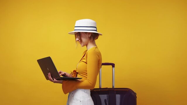 Online tickets. Excited woman. Vacation planning. Positive inspired lady in hat searching on laptop posing with suitcase on yellow background.