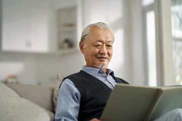 asian old man sitting on couch reading book at home