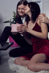 Cropped close-up shot of a dark-haired girl in a black suit hugs her beloved. Surprised woman in a red dress receiving gift from her girlfriend. Young lesbian couple in a romantic mood. LGBT concept.