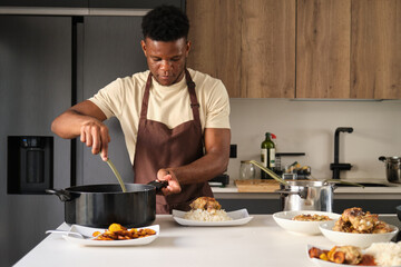 Fototapeta na wymiar Young black man serving tomato sauce on fried chicken mince and rice on a plate in a kitchen.