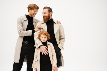 happy young man in trench coat hugging bearded father near redhead son isolated on grey.