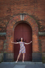 Fototapeta na wymiar A beautiful slender smiling woman with long dark hair, in a light summer dress with a floral pattern, stands at the door in the form of an arch of an old red brick building.