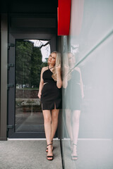 A stylish slender girl with blond long hair and long legs stands against the backdrop of a modern building. Dressed in a short black sexy skirt and top.