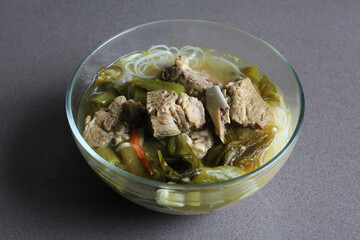 sup bakut or baikut babi sayur asin, is popular Chinese Indonesian pork rib soup with pickled cabbage or mustard greens and vermicelli in clear bowl, on black background, front view.