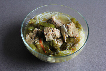 sup bakut or baikut babi sayur asin, is popular Chinese Indonesian pork rib soup with pickled cabbage or mustard greens and vermicelli in clear bowl, on black background.