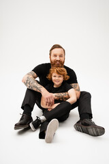 full length of tattooed man with red haired grandson sitting in black clothes on light grey background.