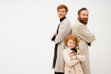 Fototapeta na wymiar joyful bearded man in trench coats standing back to back with crossed arms near redhead boy isolated on grey.
