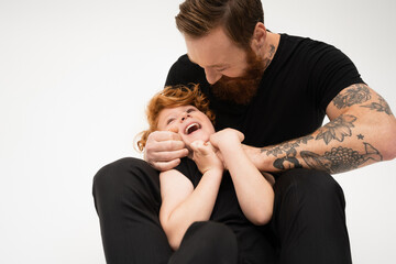 bearded and tattooed grandpa playing with excited redhead grandson isolated on grey.