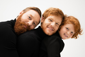 happy and bearded men with redhead boy smiling at camera while leaning on each other isolated on grey.