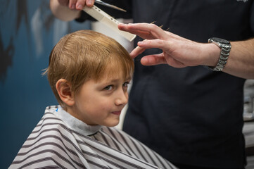 Happy cute fair-haired preschool boy getting a haircut. Children's hairdresser with scissors and comb cuts a little boy's hair in a room with a loft interior