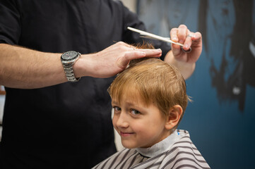 Happy cute fair-haired preschool boy getting a haircut. Children's hairdresser with scissors and comb cuts a little boy's hair in a room with a loft interior