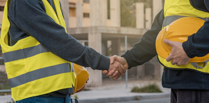 Dos operarios de construcción, estrechándose la mano y de fondo un edificio en obra.