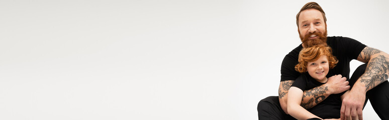 tattooed and bearded man embracing red haired grandson while sitting and smiling at camera isolated on grey, banner.