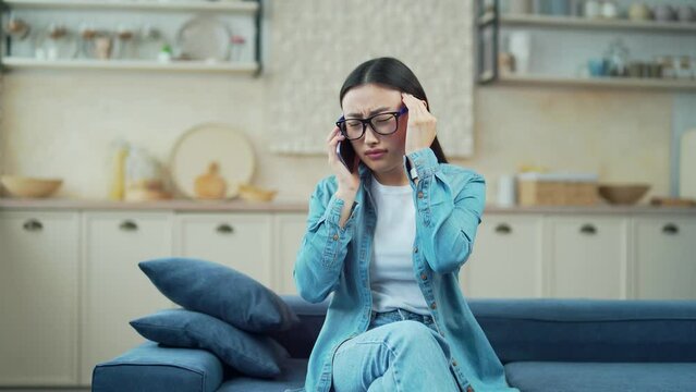Young asian woman having a headache talking on the phone while sitting on the sofa in the living room. A tired female in glasses and casual clothes gives head massage while talking on a smartphone