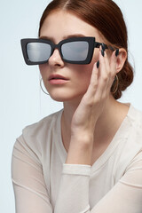 Naklejka premium Cropped close-up shot of a young woman wearing mirrored sunglasses with gray lenses. A girl holding her rectangular sunglasses with wide temples and a black frame is on a light background. Front view.
