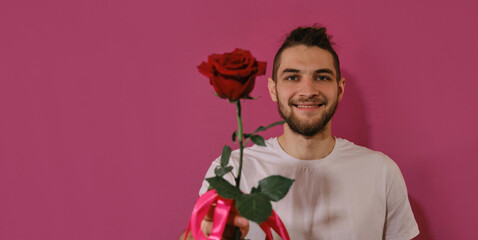 Concept of Valentine's Day. International Women's Day greeting card. Young Caucasian man presents one red rose stretching forward and smiling. Isolated on pink background. Web banner.