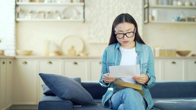 Young asian woman reading a letter with bad news while sitting on the sofa in the living room. Sad female opens envelope, she is disappointed by unpleasant information, getting dismissal notice